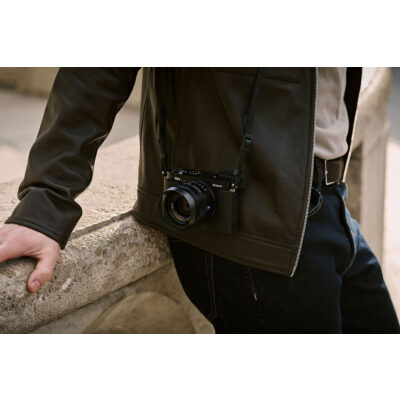 Sony DSC RX1R III hangt aan een schouderband, steunt op een muur terwijl het een casual outfit complementeert.