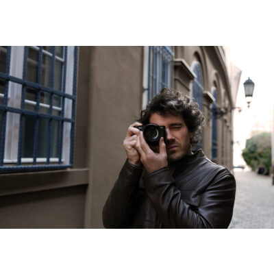 Sony DSC RX1R III in handen van een man, fotograferend tegen een historische achtergrond met ramen en straatverlichting.