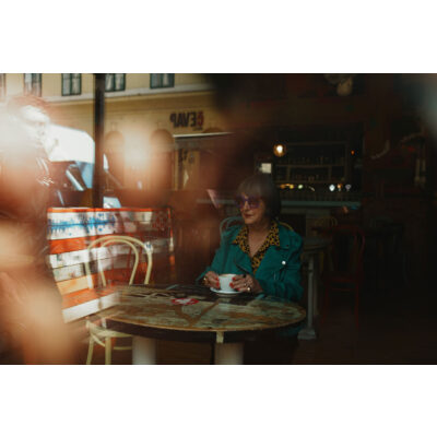 Sony DSC RX1R III toont een vrouw met grijze haren, in een groene leren jas en paarse zonnebril, die koffie drinkt aan een kleurrijke tafel.